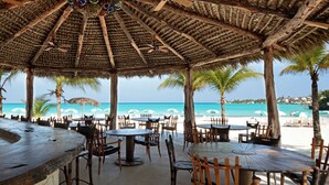 An outdoor dining area with thatched umbrellas and wooden tables, overlooking a beach with palm trees and the ocean. - The Island House (Nassau)