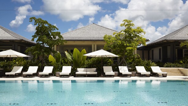 A pool area with white lounge chairs and umbrellas, surrounded by greenery and buildings with grey roofs. - The Island House (Nassau)