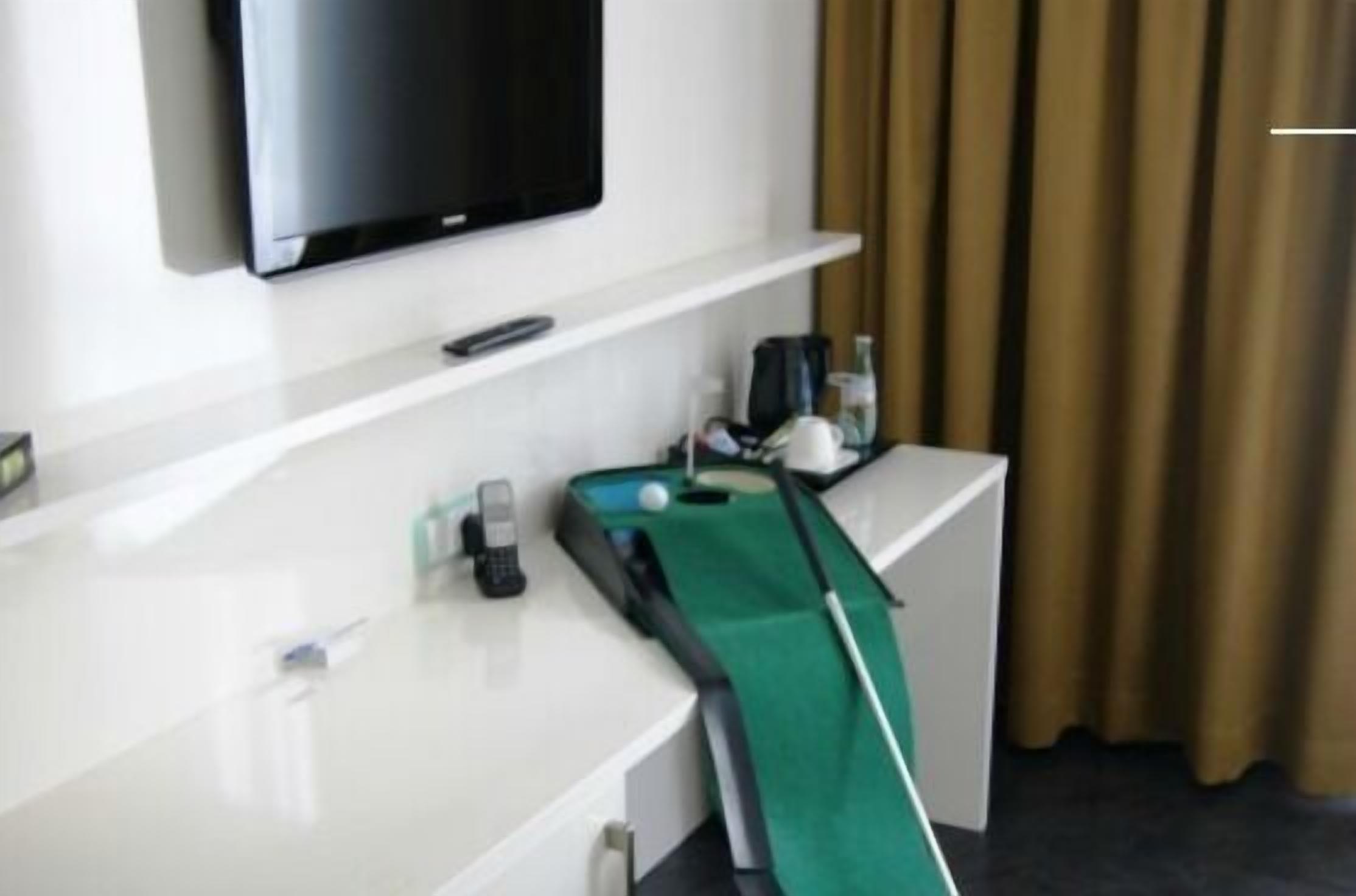 Habitación de hotel con televisor de pantalla plana en la pared, un escritorio con teléfono, mantel verde y taco de billar.