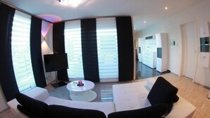 Ett modernt vardagsrum med en vit soffa, ett soffbord och en tv. Det finns ett stort fönster med persienner och en taklampa.