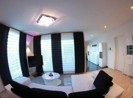 Una sala de estar moderna con un sofá blanco, una mesa de centro y un televisor. Hay una ventana grande con persianas y una lámpara de techo.