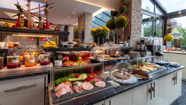 A buffet with various food items, including meats, fruits, and vegetables, in a modern kitchen setting with large windows and decorative elements. - Hotel Zum Rosengarten (Darmstadt)