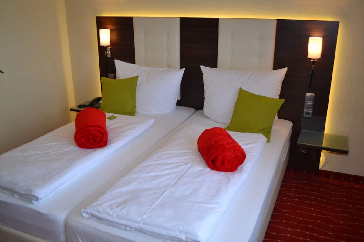 A hotel room with two single beds, each with a white mattress and pillows, and red towels. There is a nightstand with a lamp and a phone.