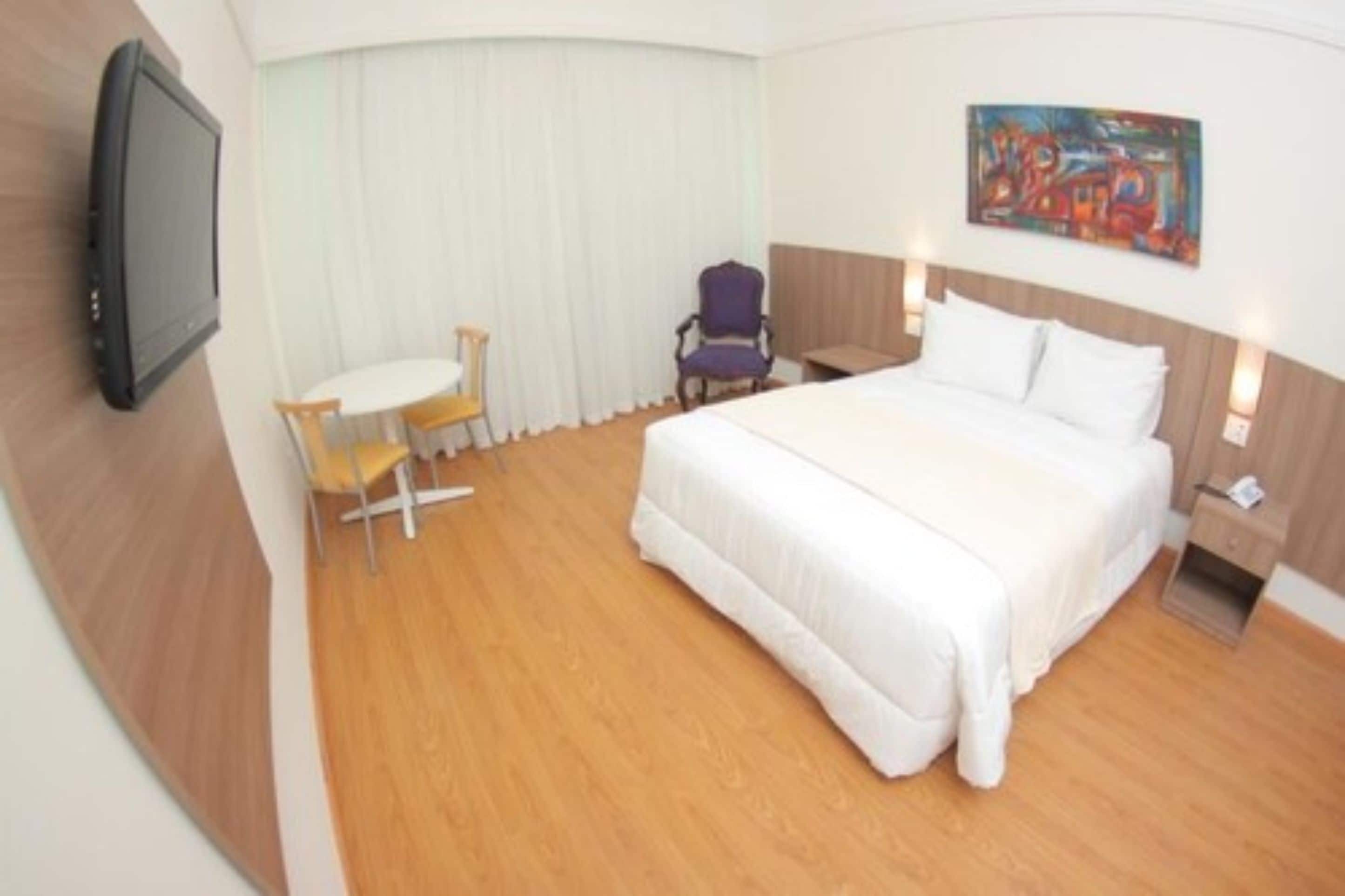 Ein Hotelzimmer mit einem Bett, einem Fernseher, einem kleinen Tisch mit Stühlen und einem Bild an der Wand.