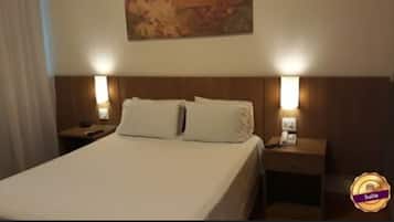 Quarto de hotel com uma cama grande, duas mesas de cabeceira, um criado-mudo e um quadro na parede.