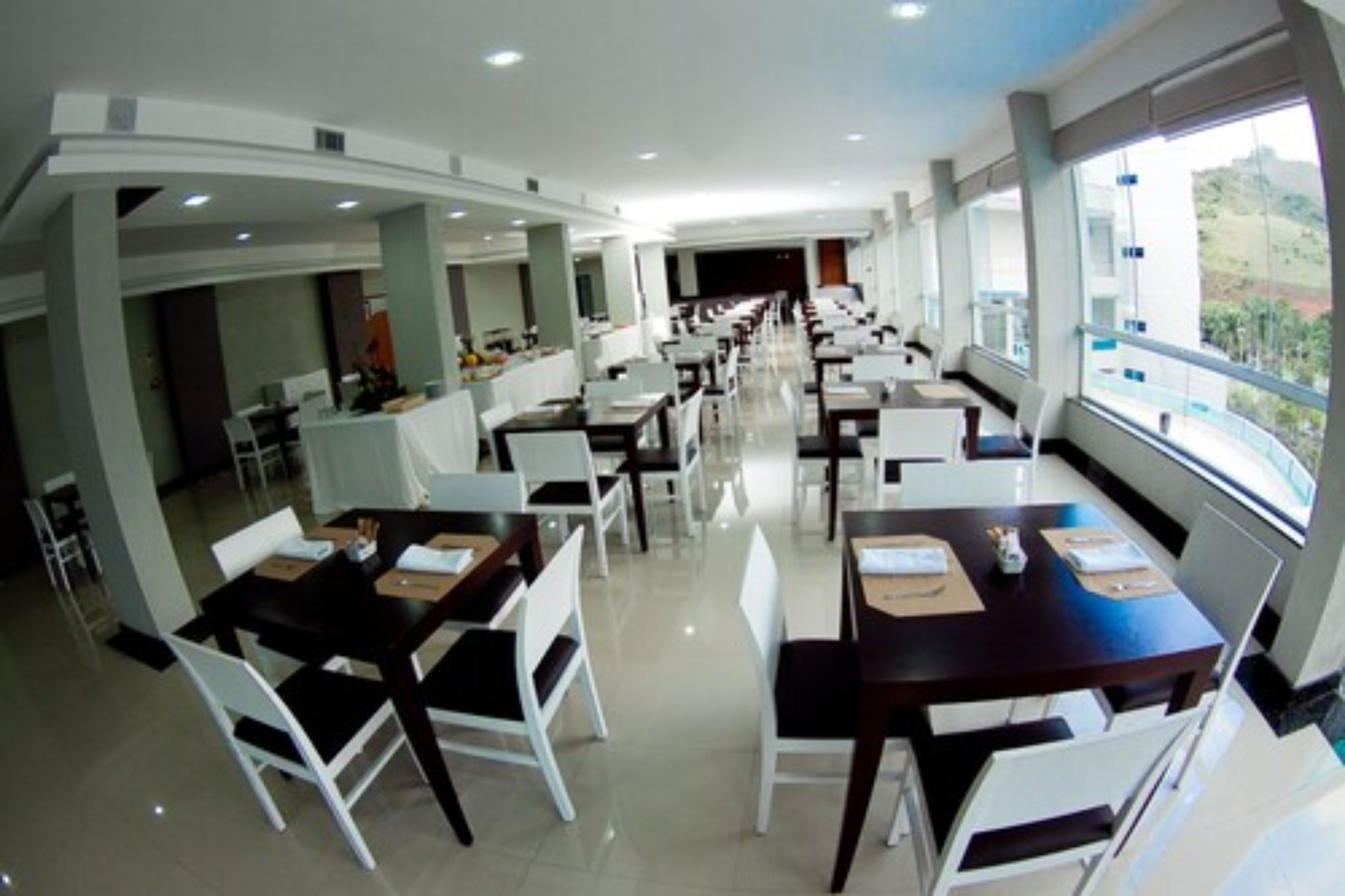 Uma sala de jantar moderna com cadeiras brancas e mesas escuras, grandes janelas com vista e um interior limpo e bem iluminado.