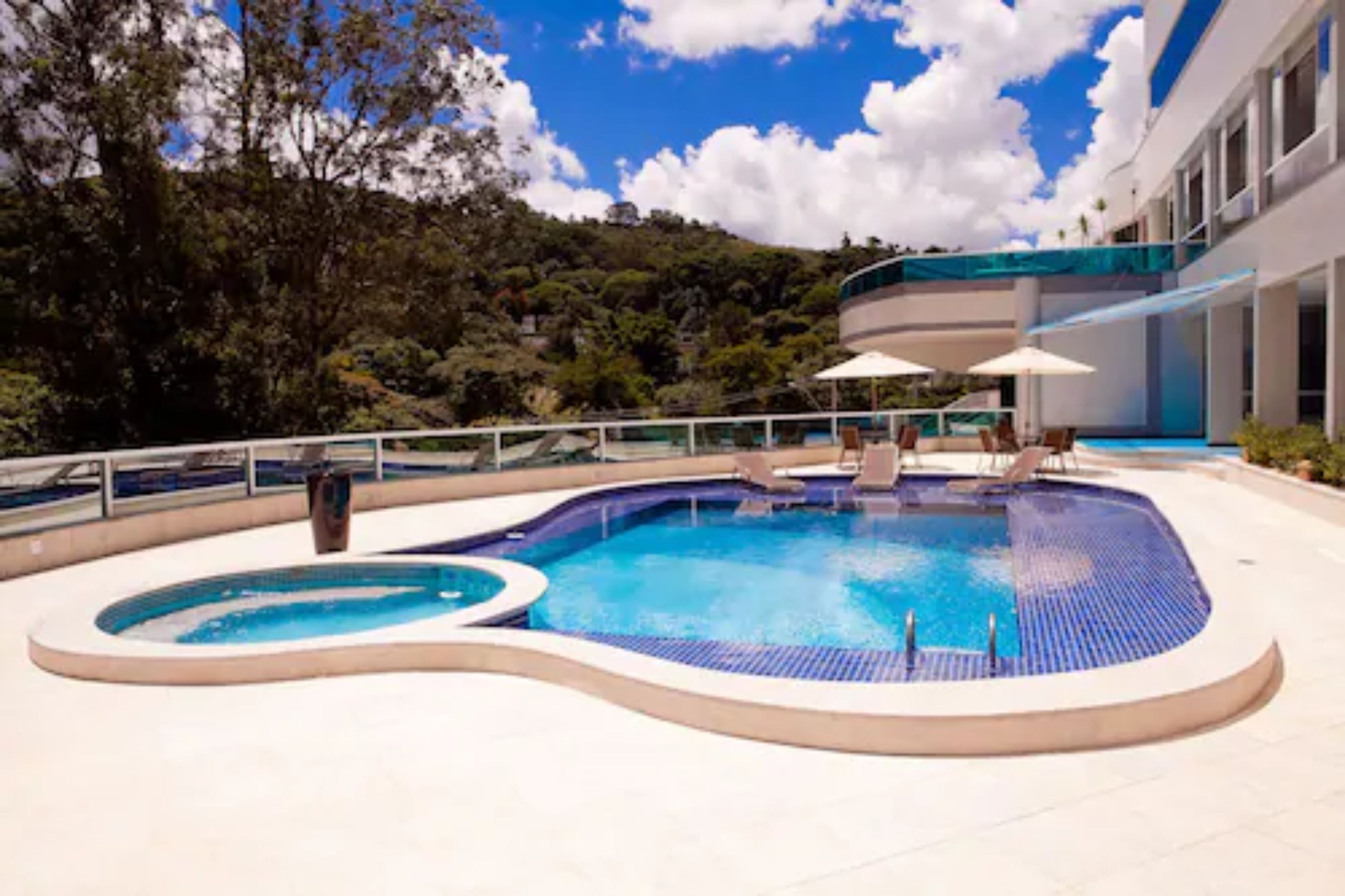 A swimming pool with a curved design, surrounded by lounge chairs and umbrellas, set against a backdrop of trees and a clear sky.