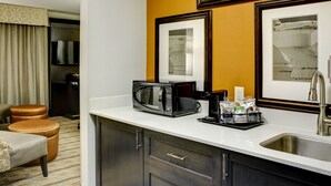 A modern hotel kitchen with a microwave, coffee maker, and sink. There are framed pictures on the wall and a seating area with a chair and ottoman. - Hampton Inn & Suites Columbus (Columbus)