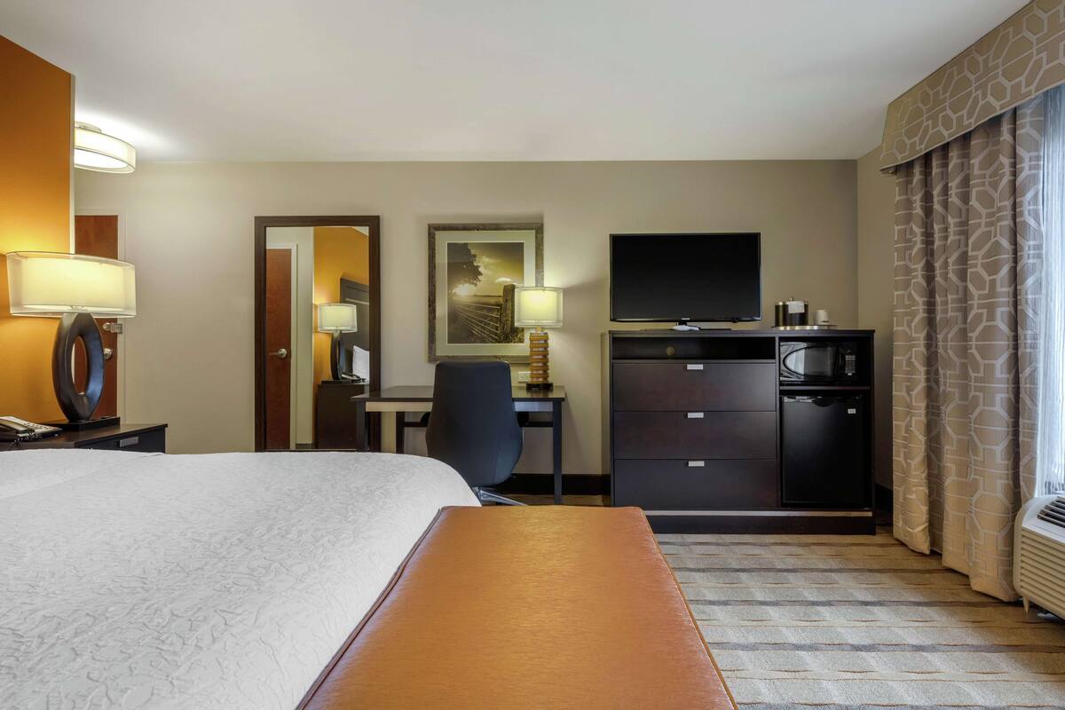 A hotel room with a large bed, a desk with a chair, a television, a minibar, and a window with curtains.
