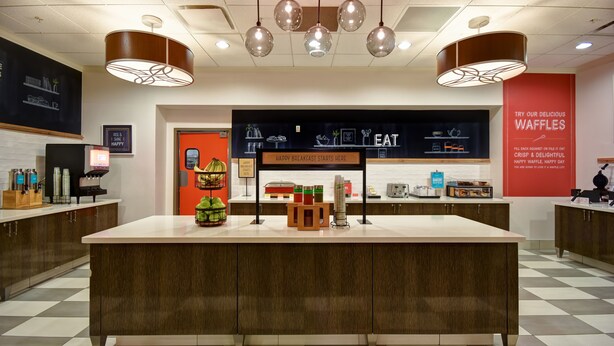 A modern cafe interior with a central island, pendant lighting, and a red accent wall. - Hampton Inn & Suites Columbus (Columbus)