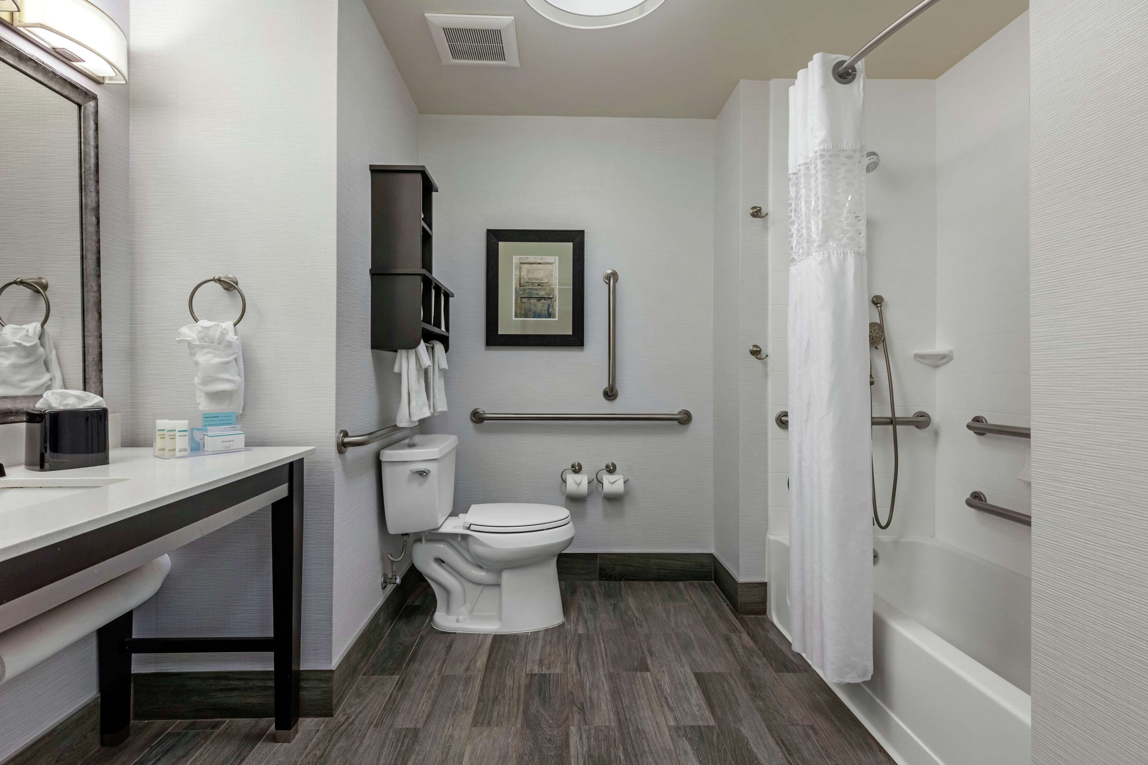 Un baño moderno con inodoro, bidé y ducha. Se aprecia un perchero, un cuadro enmarcado en la pared y una mesita con artículos de aseo.