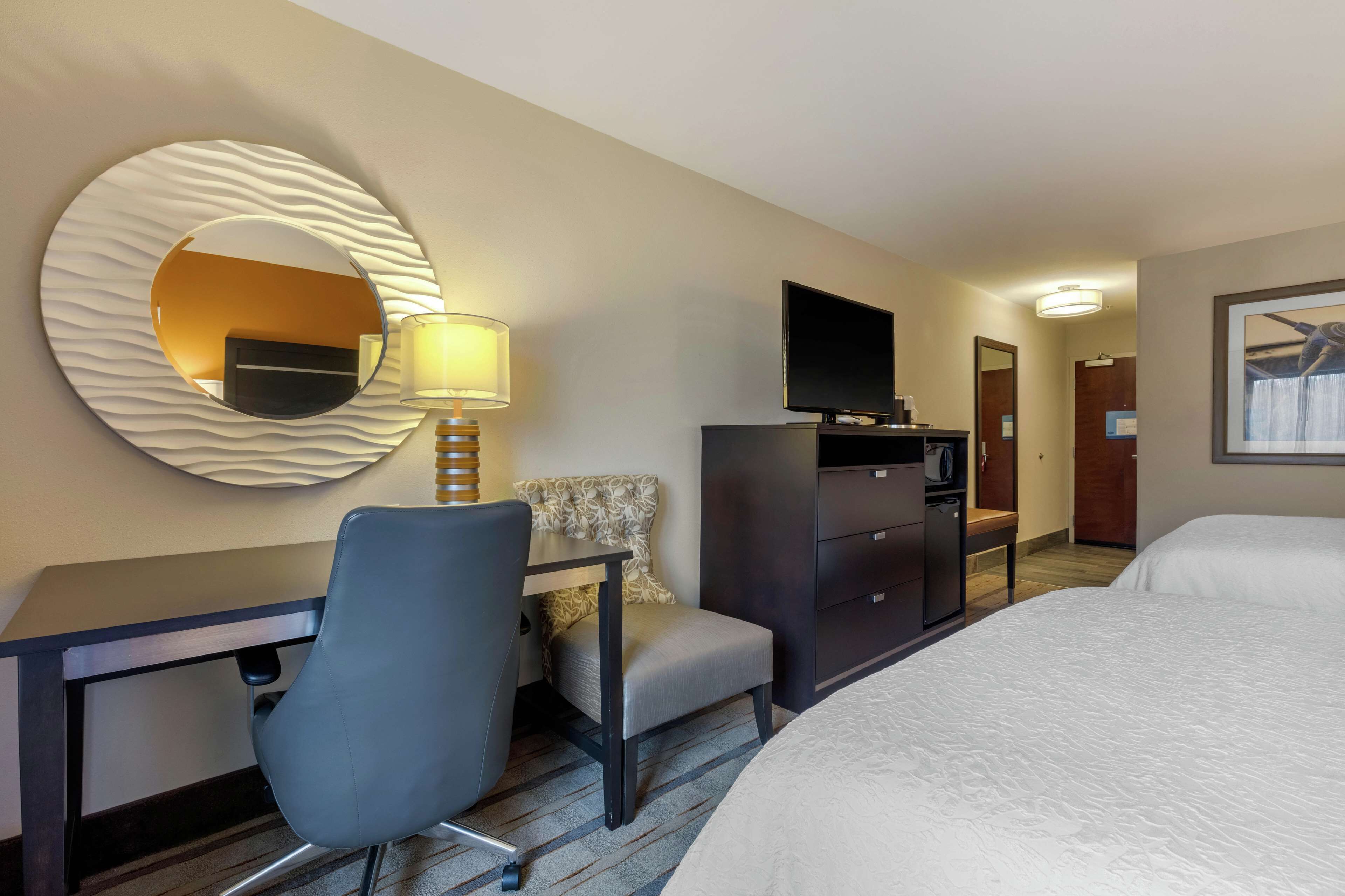 A hotel room with a bed, a desk with a chair, a television, a mirror, and a lamp.