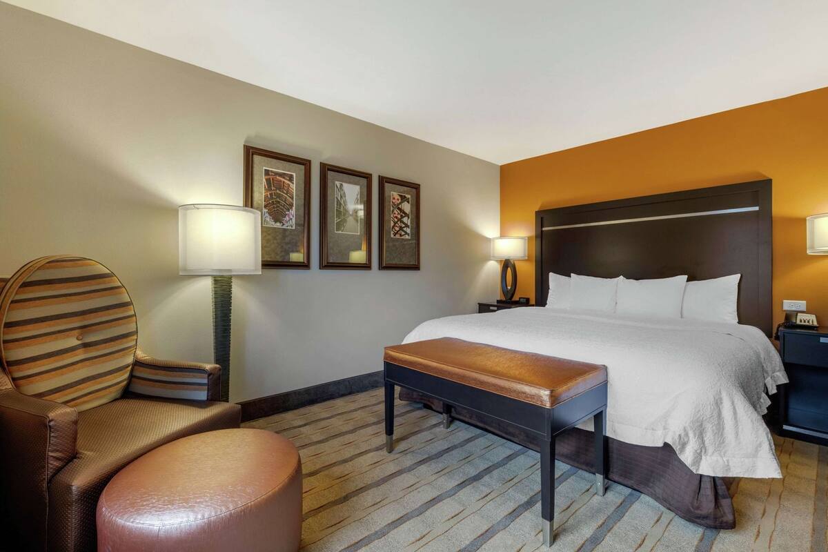 A hotel room with a bed, a chair, a bench, a nightstand, a lamp, and framed pictures on the wall.