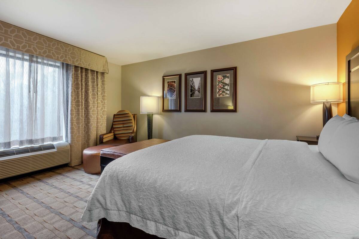 A hotel room with a large bed, a chair, a desk, and a window with curtains.