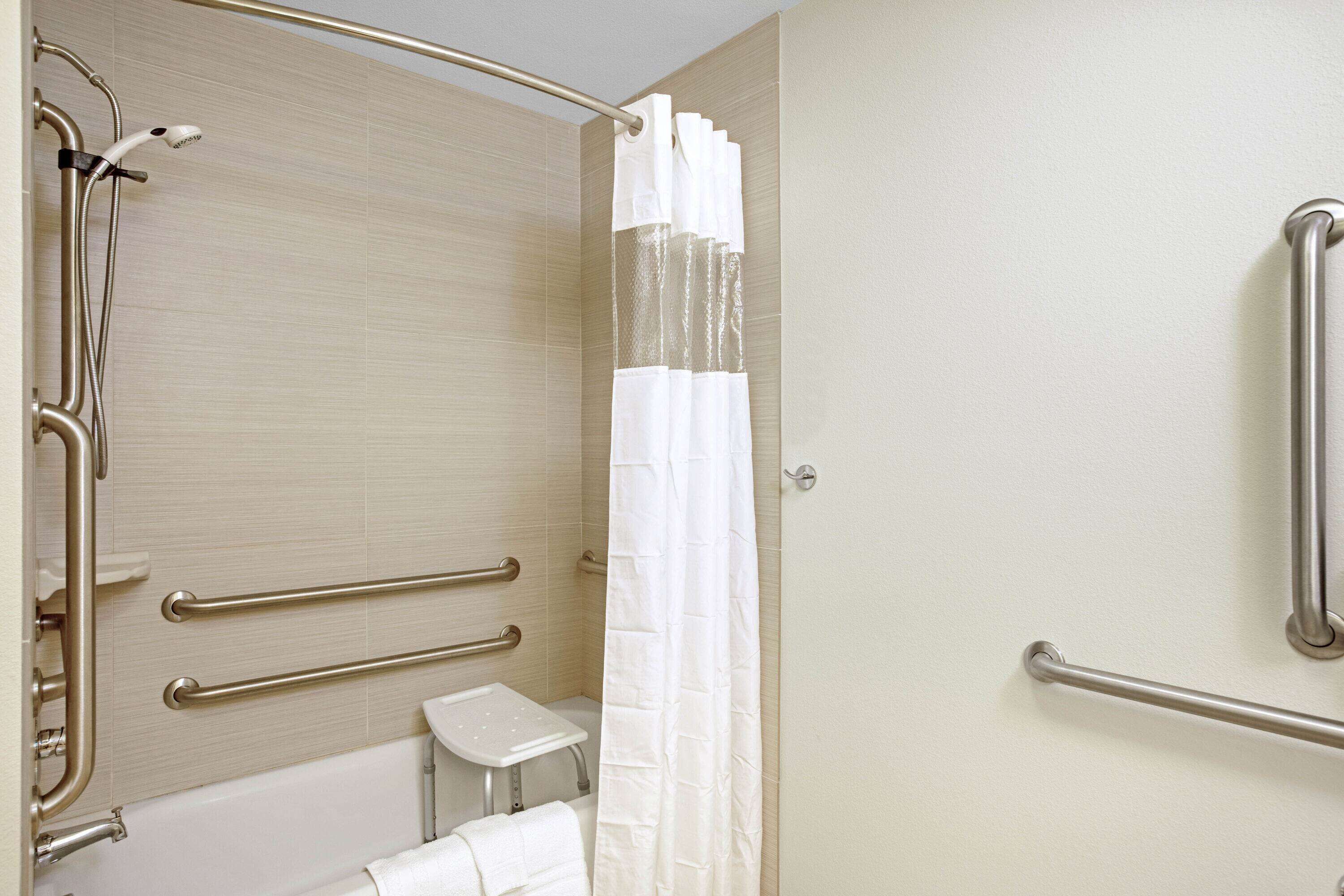 Un baño con bañera, ducha con cabezal de ducha manual y cortina de ducha blanca. Hay barras de apoyo en las paredes y un taburete pequeño cerca de la bañera.
