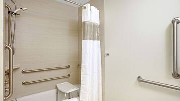 Un baño con bañera, ducha con cabezal de ducha manual y cortina de ducha blanca. Hay barras de apoyo en las paredes y un taburete pequeño cerca de la bañera.