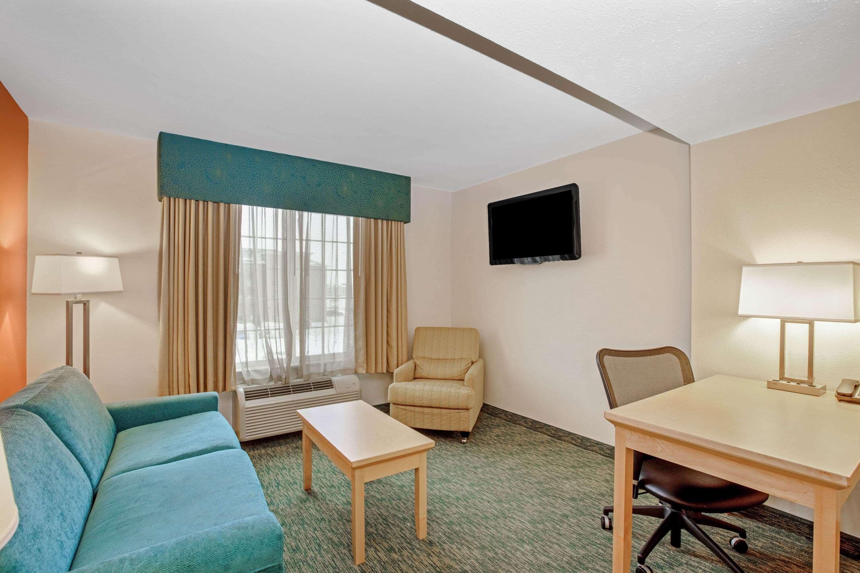 A hotel room with a sofa, a small table, a chair, a TV mounted on the wall, and a window with curtains.