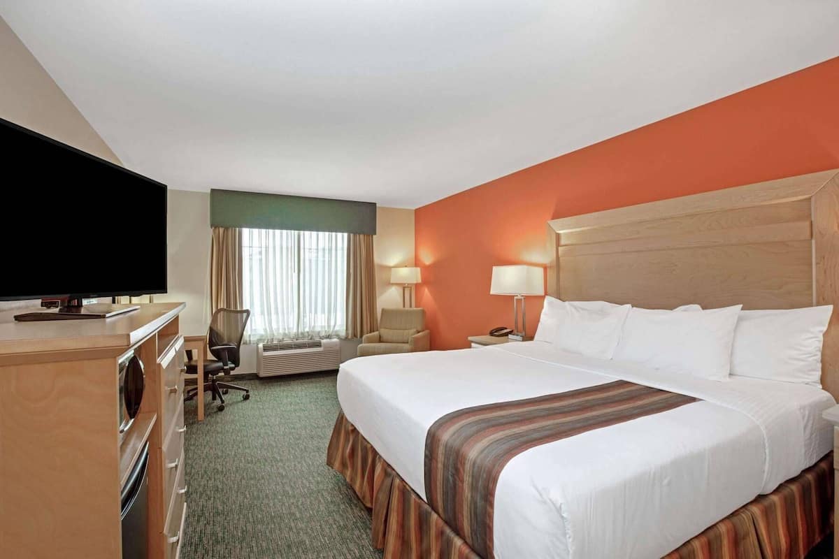 Habitación de hotel con cama, escritorio, televisor, silla, lámpara y ventana con cortinas.