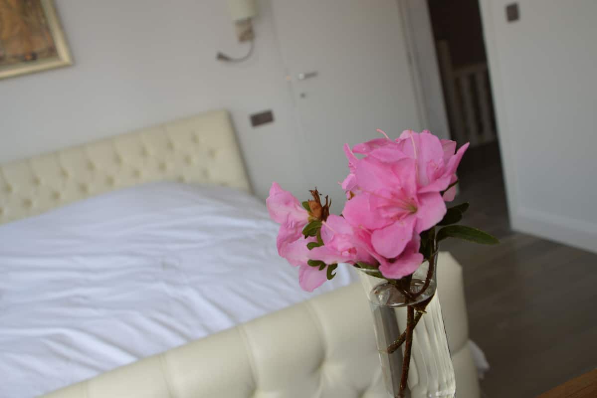 Un dormitorio con una cama blanca y un florero con flores rosas sobre la mesita de noche.