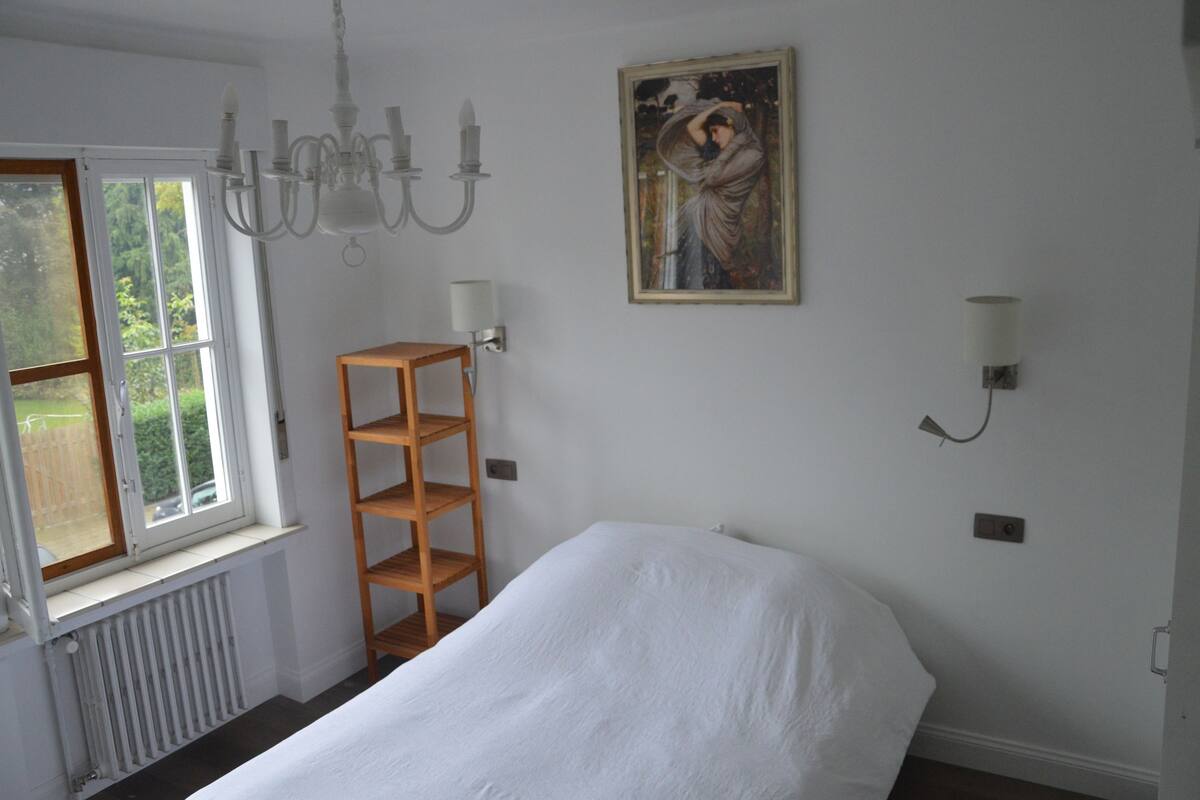 Un dormitorio con cama, ventana, radiador, estante de madera y un cuadro en la pared.