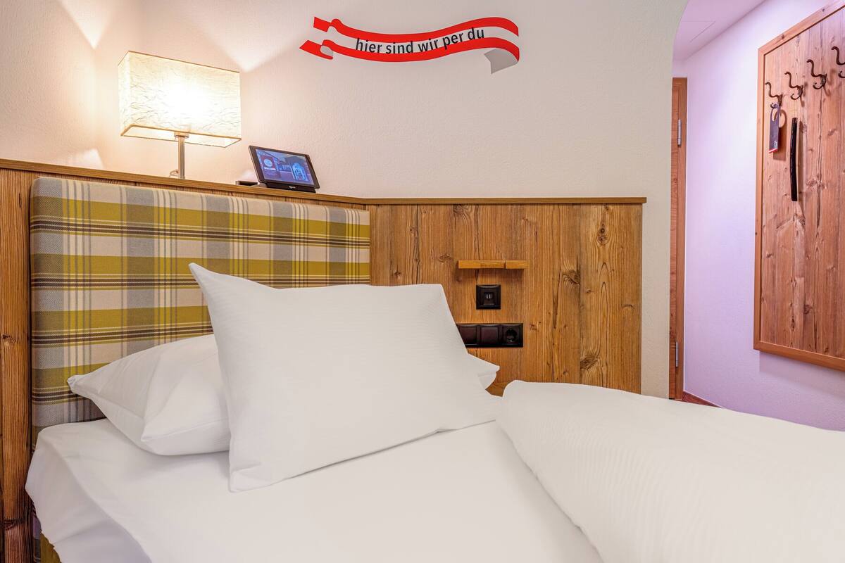 Quarto de hotel com cabeceira xadrez, luminária de cabeceira, mesinha com um laptop e um aviso na parede.