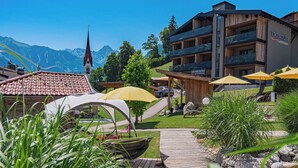 Un hotel con fachada de madera, mobiliario exterior con sombrillas amarillas y vistas a la montaña.