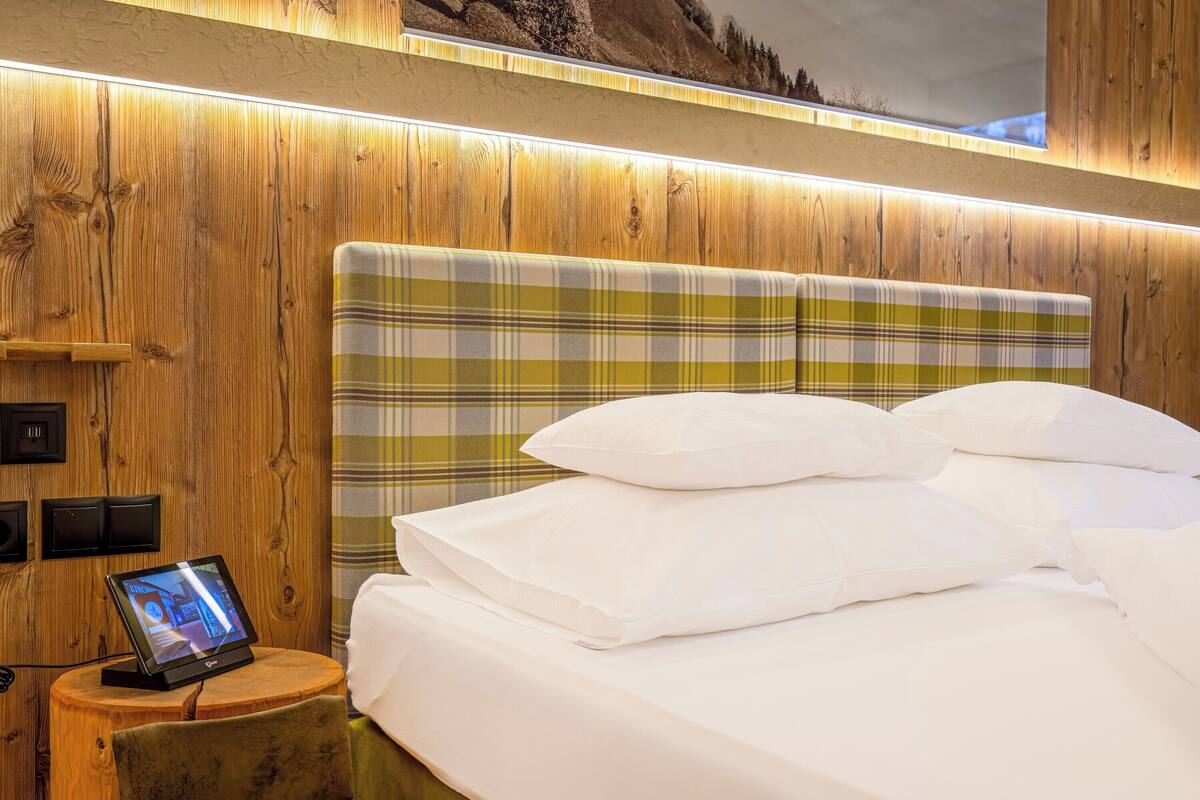 Quarto de hotel com cabeceira de madeira, mesa de cabeceira com relógio digital e cama com roupa de cama branca.