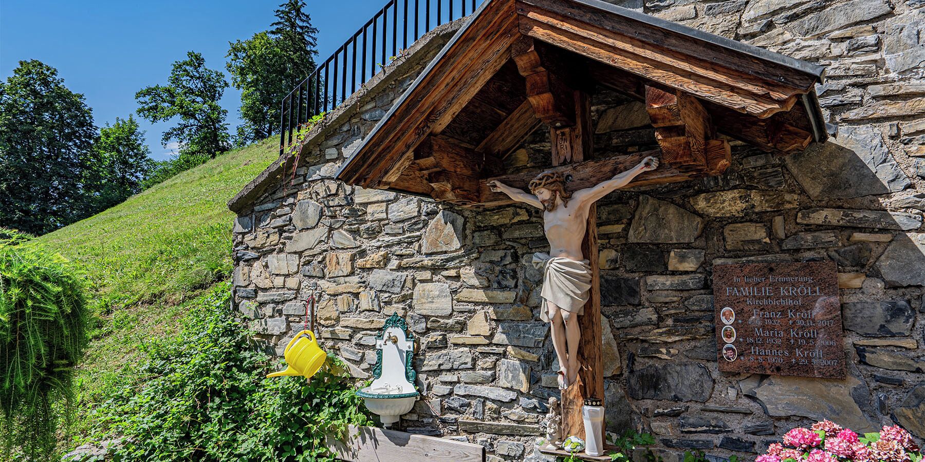Una capilla de piedra con una estatua de la crucifixión, una lápida con nombres y un cubo amarillo.