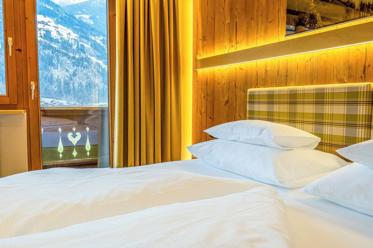 Quarto com cabeceira de madeira, cama com roupa de cama branca e vista para montanhas cobertas de neve pela janela.