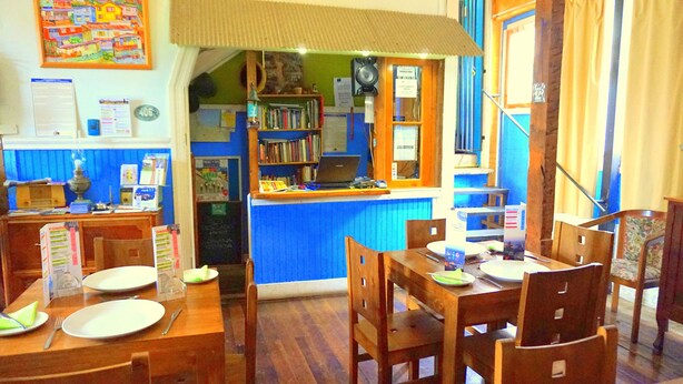 Ein Restaurant mit Holztischen und -stühlen, einer Theke mit einem Laptop und einem Bücherregal im Hintergrund.