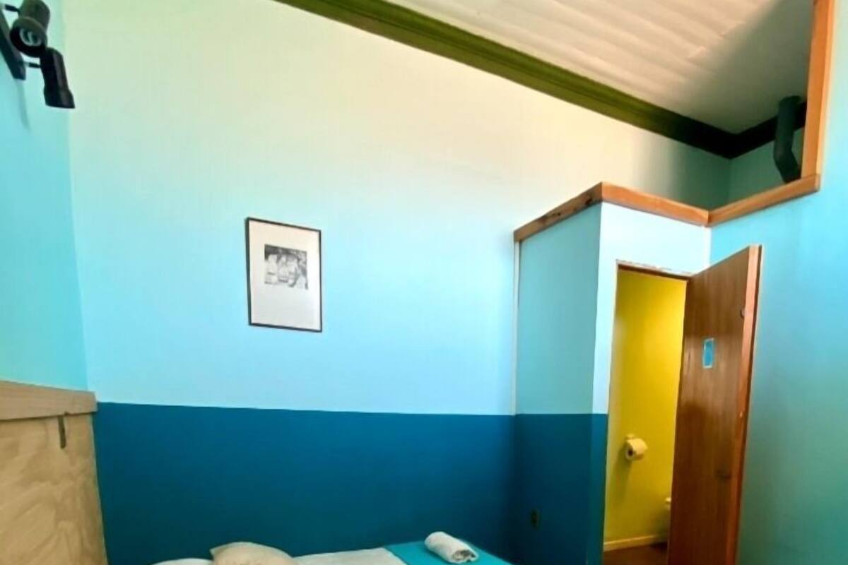 A room with two beds, each with a beige quilt and pillows, a blue wall, and a wooden door leading to a bathroom.