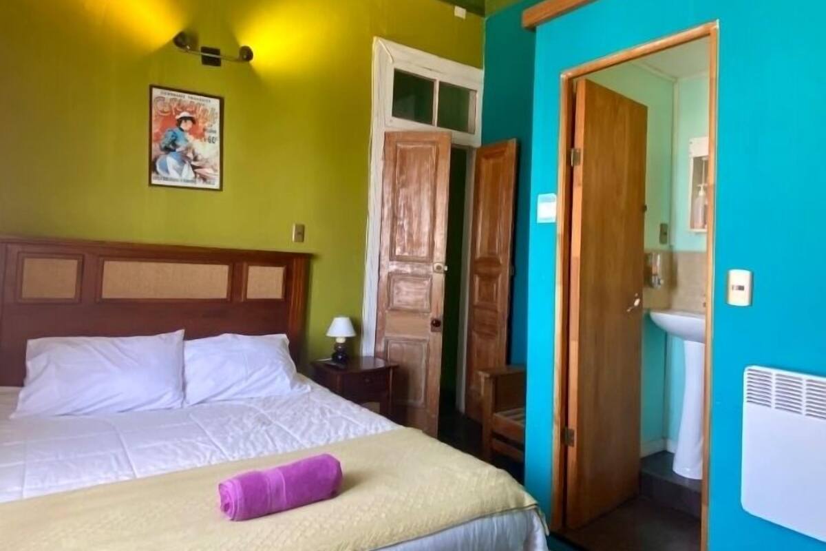 A room with a bed, a wooden wardrobe, a bathroom with a mirror, and a wall with a poster.