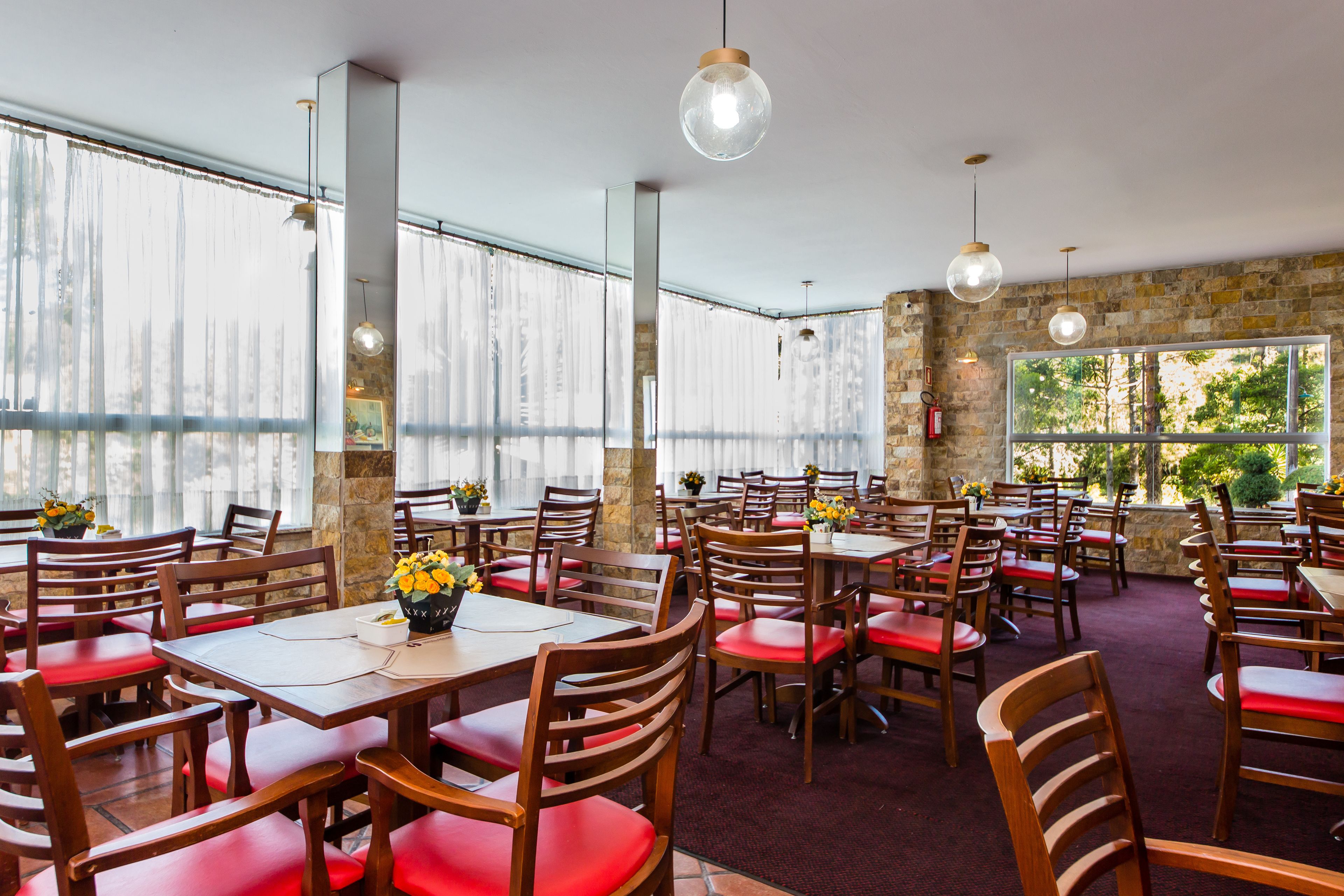 A restaurant with multiple tables and chairs, large windows with curtains, and hanging pendant lights.