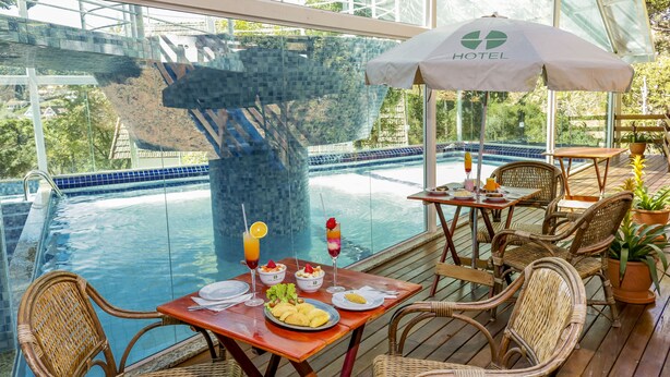 An outdoor dining area with wicker chairs and tables, overlooking a swimming pool with a glass bridge in the background.