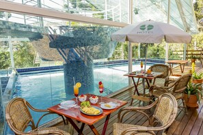 An outdoor dining area with wicker chairs and tables, overlooking a swimming pool with a glass bridge in the background. - Hotel Fazenda Golden Park Campos do Jordão (Campos do Jordão)