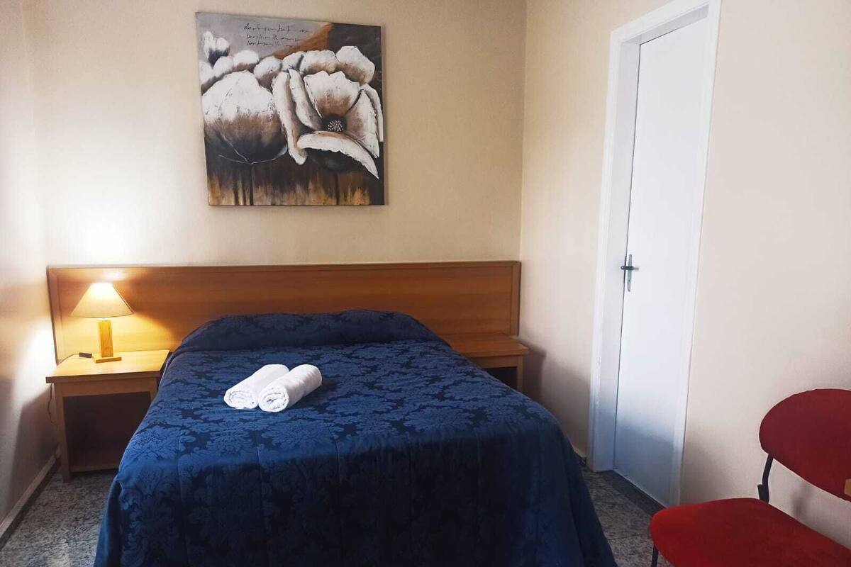 A hotel room with a bed, a nightstand with a lamp, a painting on the wall, a door, and a red chair.