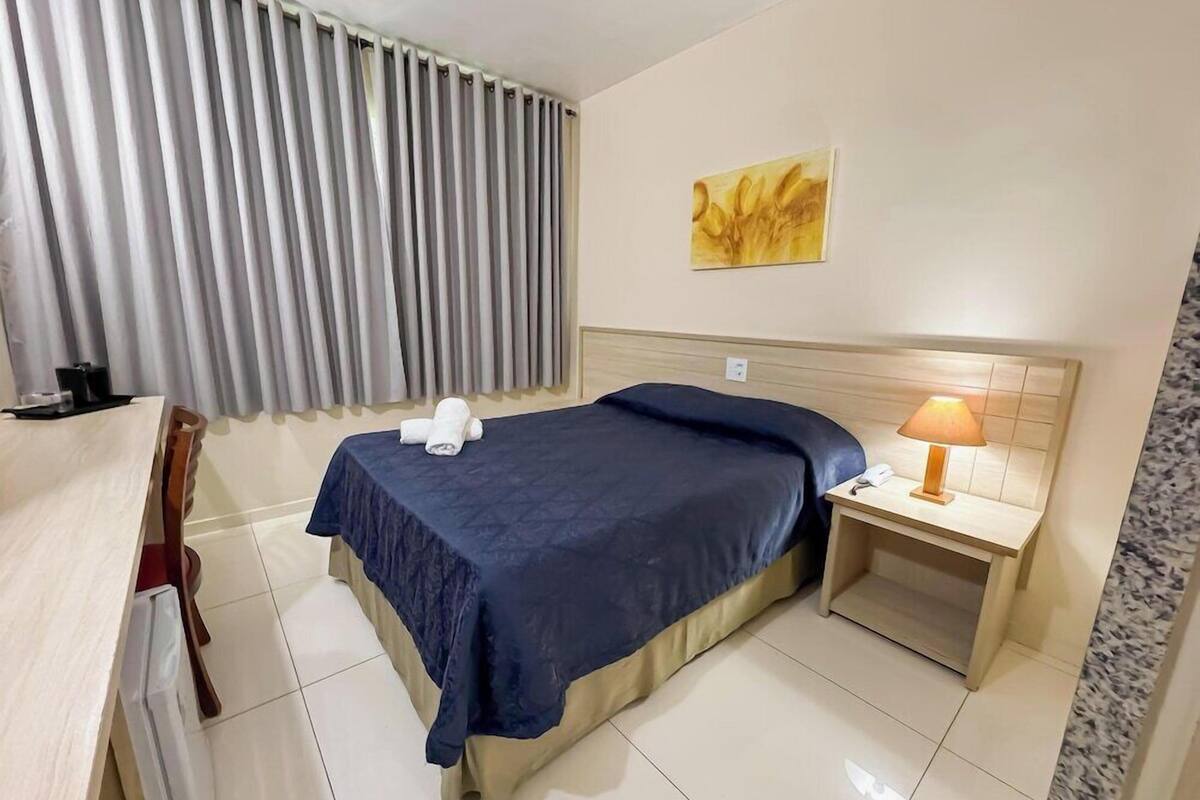 A hotel room with a bed, a bedside table with a lamp, a chair, a desk, and a window with curtains.