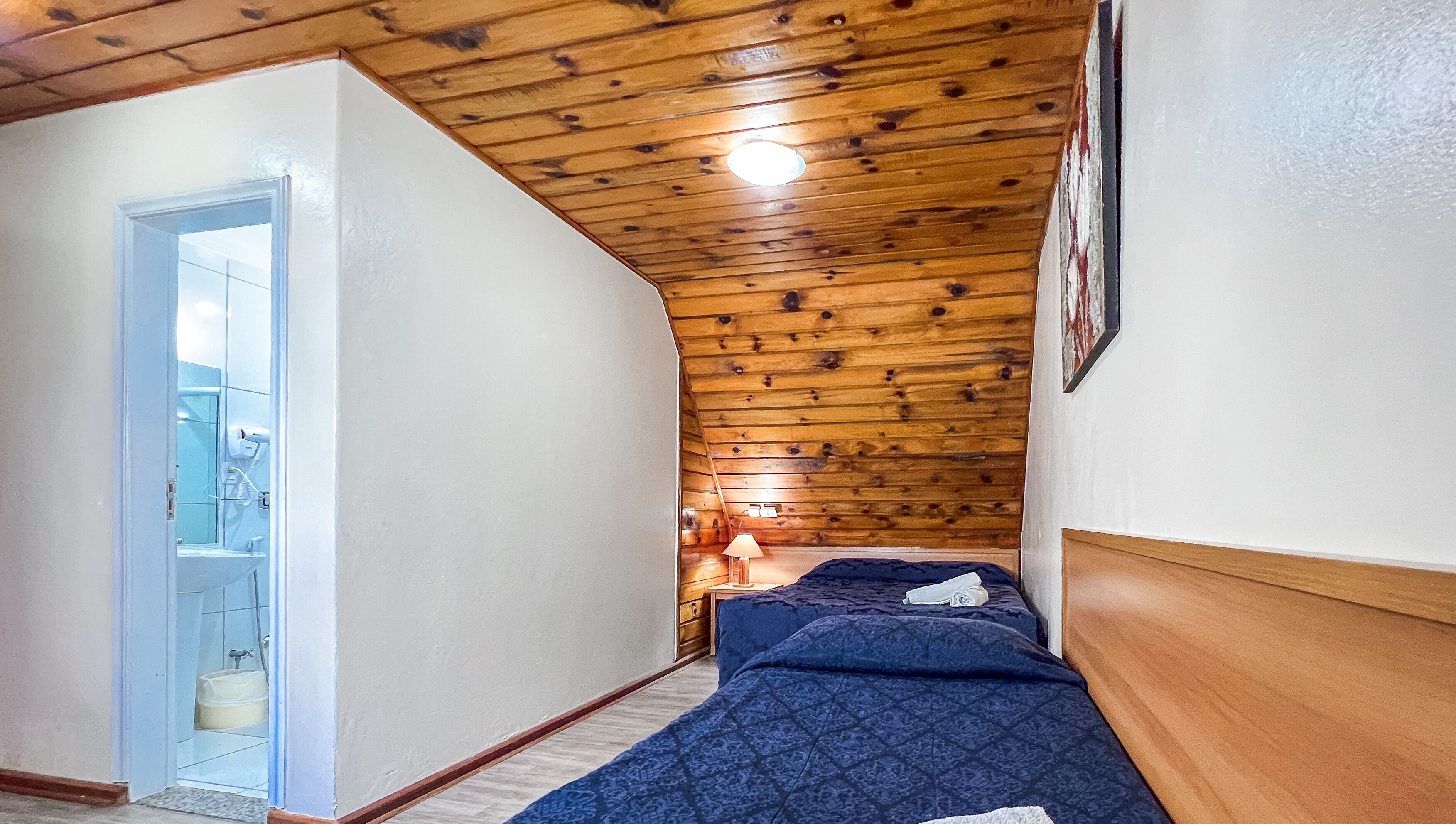 Een smalle gang die leidt naar een slaapkamer met een houten plafond, een bed en een nachtkastje met een lamp.