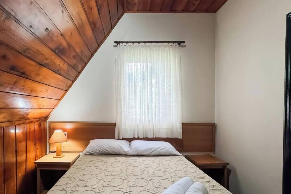 A bedroom with a sloped ceiling, wooden walls, a bed with a patterned bedspread, a bedside lamp, and a window with sheer curtains.