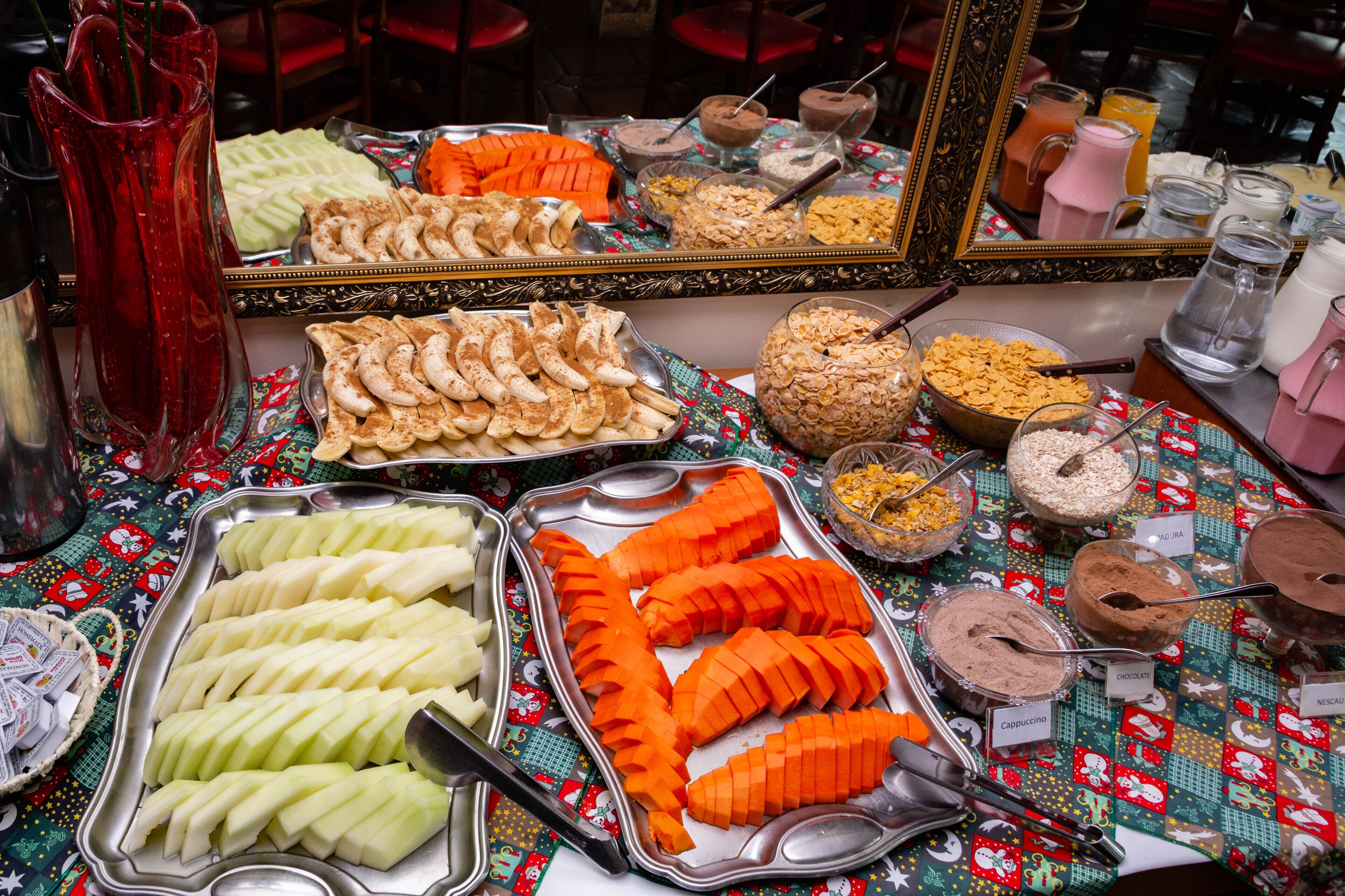Ein Buffet mit verschiedenen Speisen und Getränken, darunter Obstscheiben, Müsli und beschriftete Getränkebehälter.