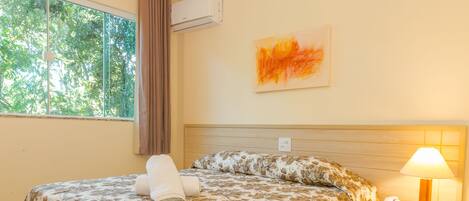 Quarto de hotel com cama, mesa de cabeceira, luminária, ar condicionado, janela com cortinas e um quadro na parede.