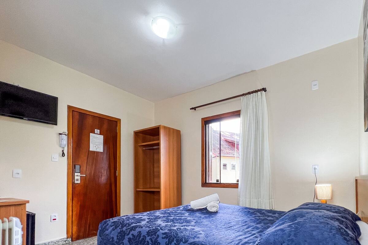 A hotel room with a bed, a desk, a telephone, a window with curtains, and a painting on the wall.