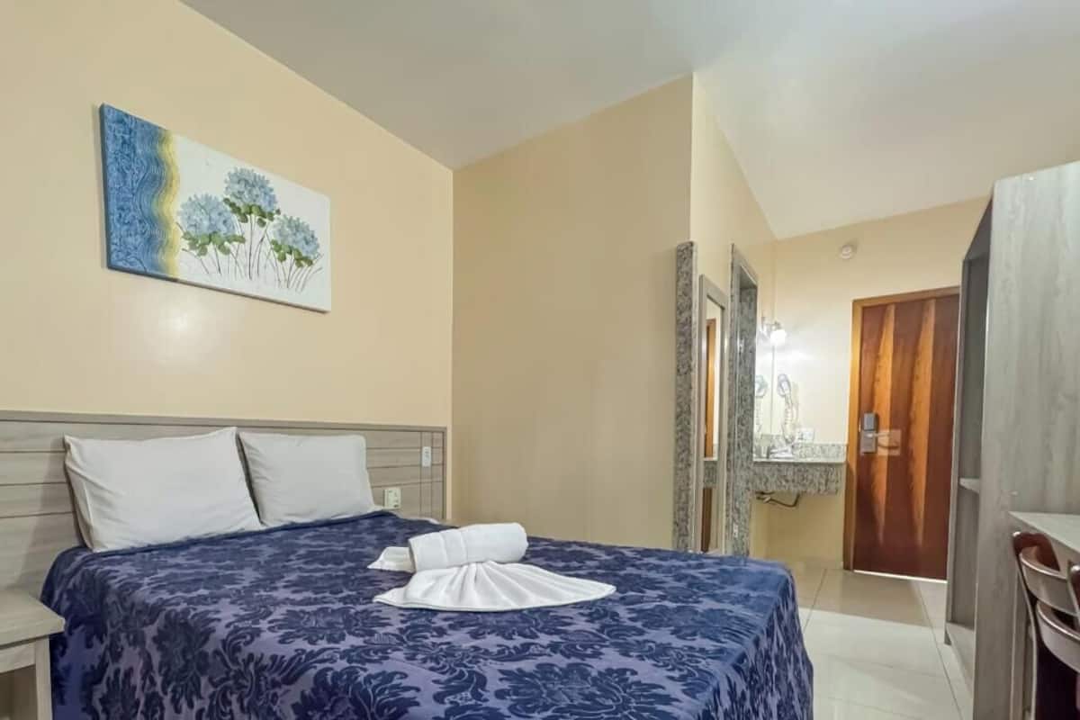 A neatly made bed with a blue floral bedspread and a white towel, a wooden headboard, a painting of flowers on the wall, and a wooden door with a mirror and light fixture.