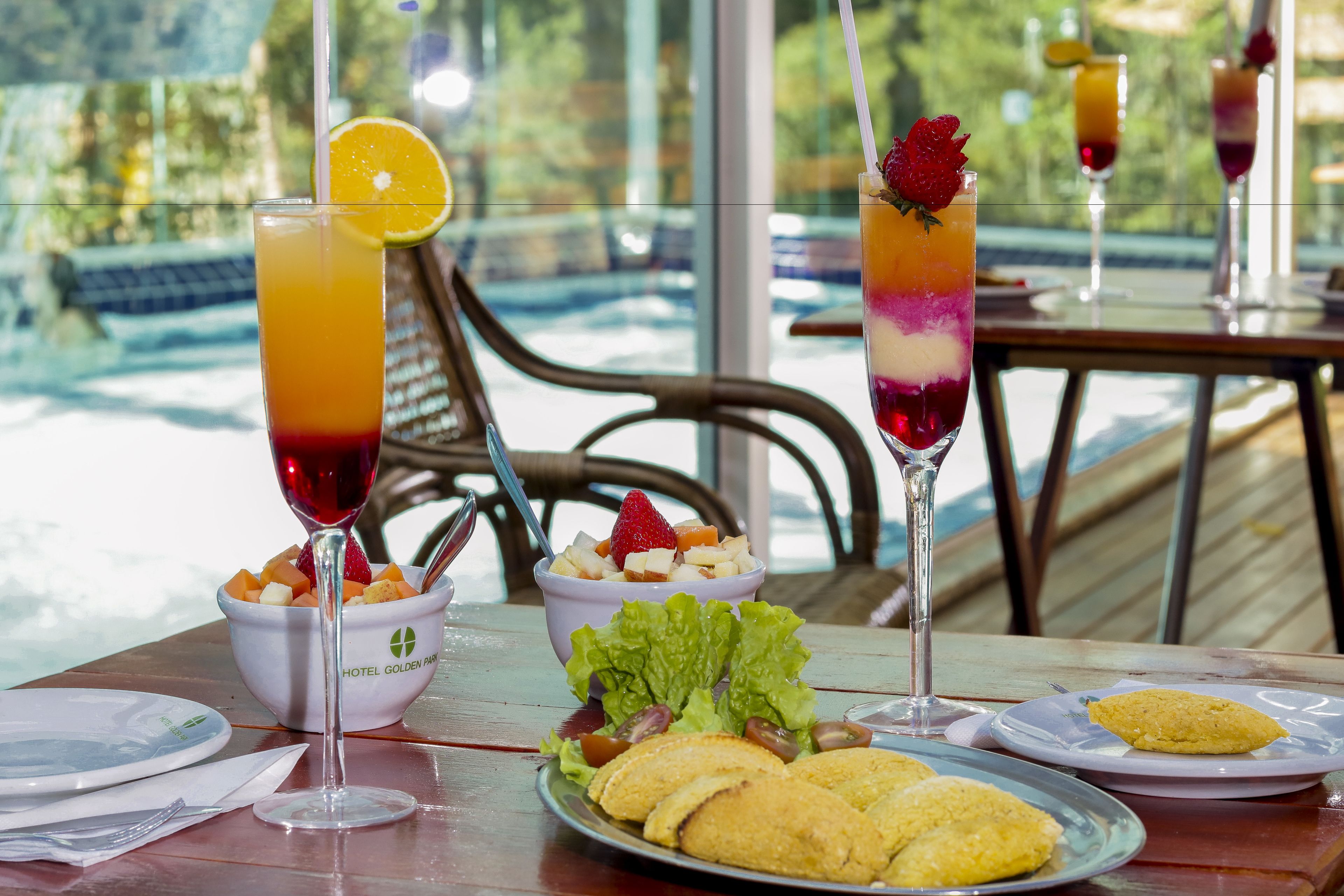 A dining table with a fruit bowl, two layered drinks, a plate of cookies, and a glass of red wine.