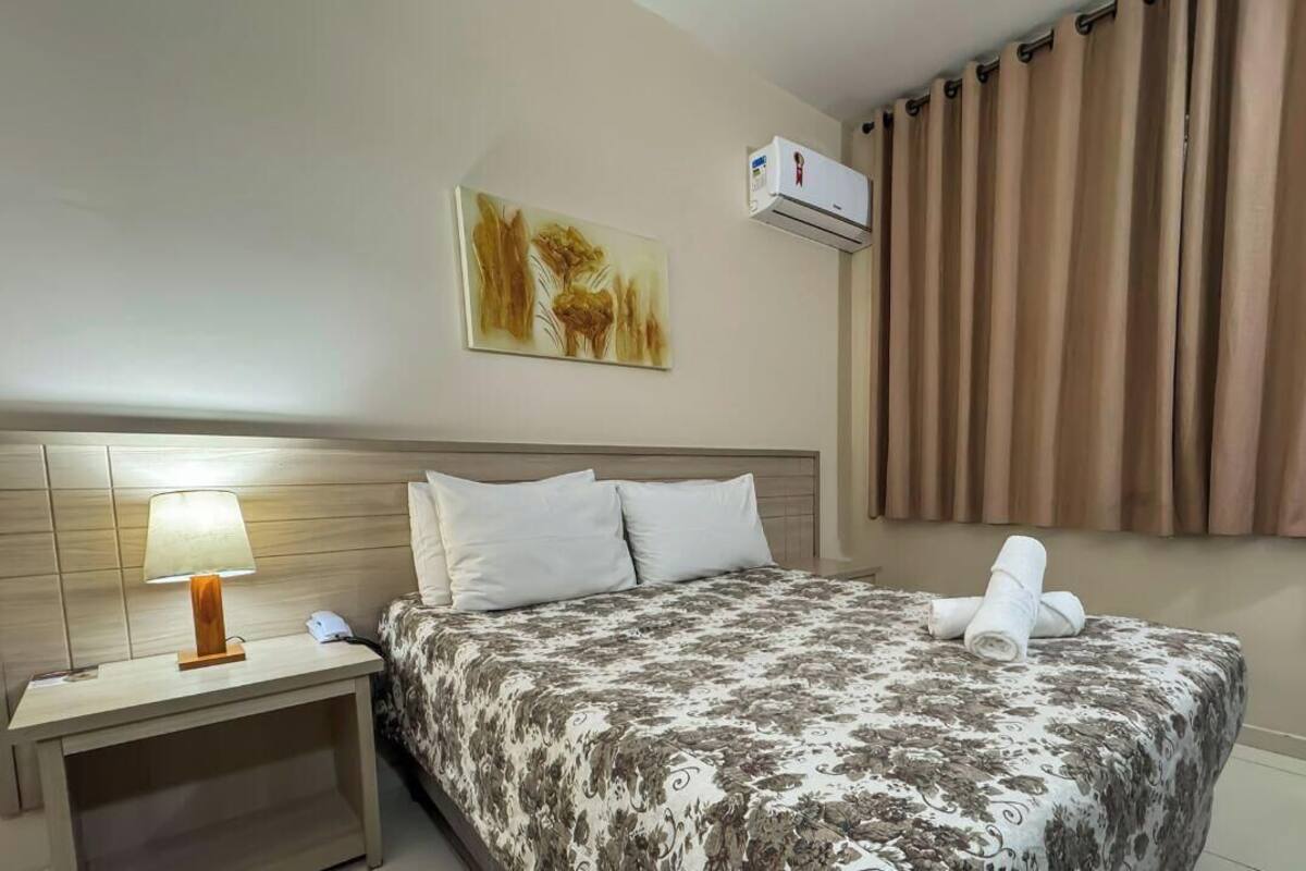A hotel room with a bed, a nightstand with a lamp, a wall-mounted air conditioner, and a window with curtains.