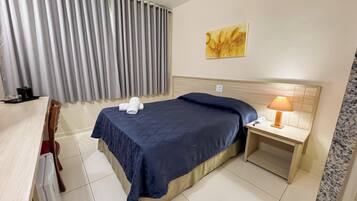A hotel room with a bed, a nightstand with a lamp, a painting on the wall, and a window with curtains.