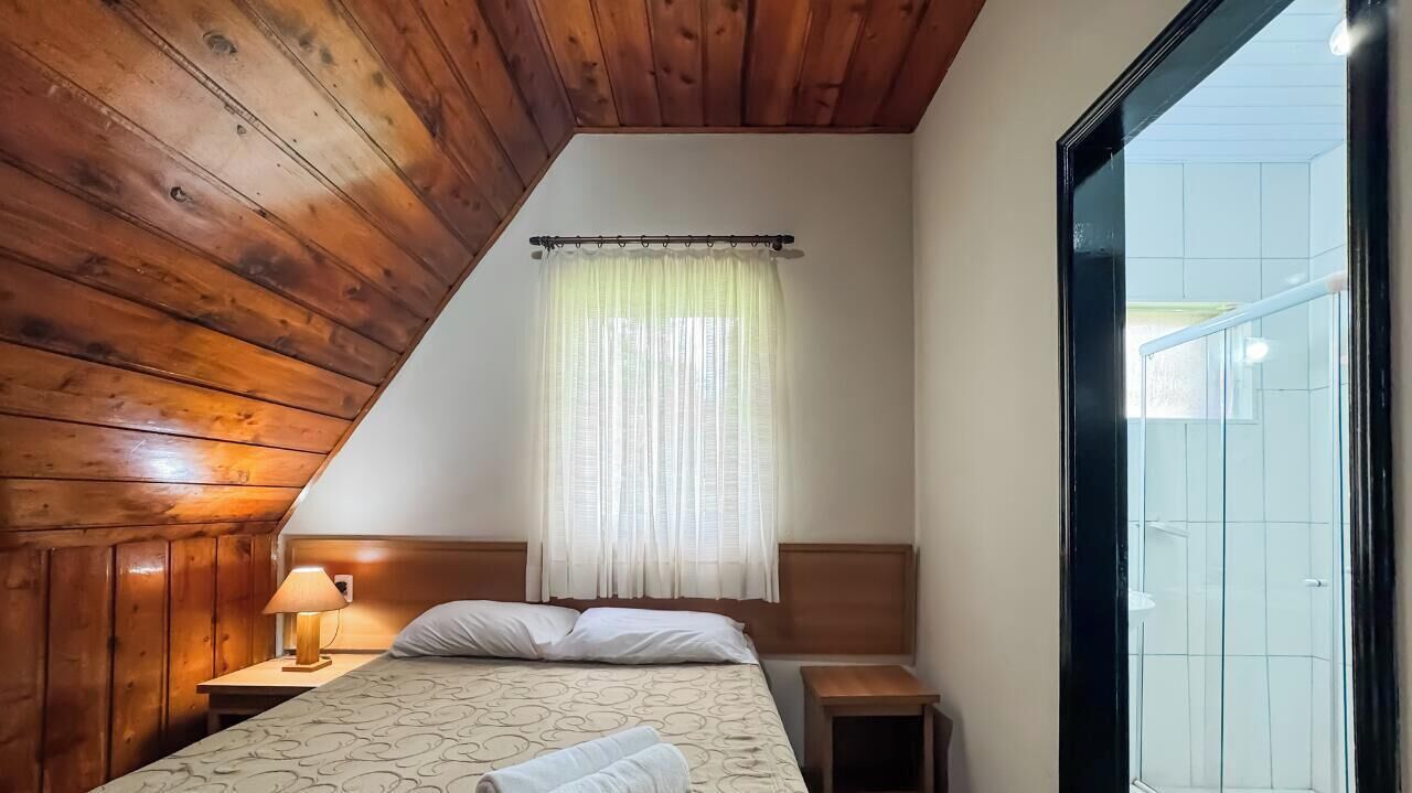 Et soveværelse med skråvæg, seng med mønstret dyne, natbord med lampe og et vindue med let gardin.