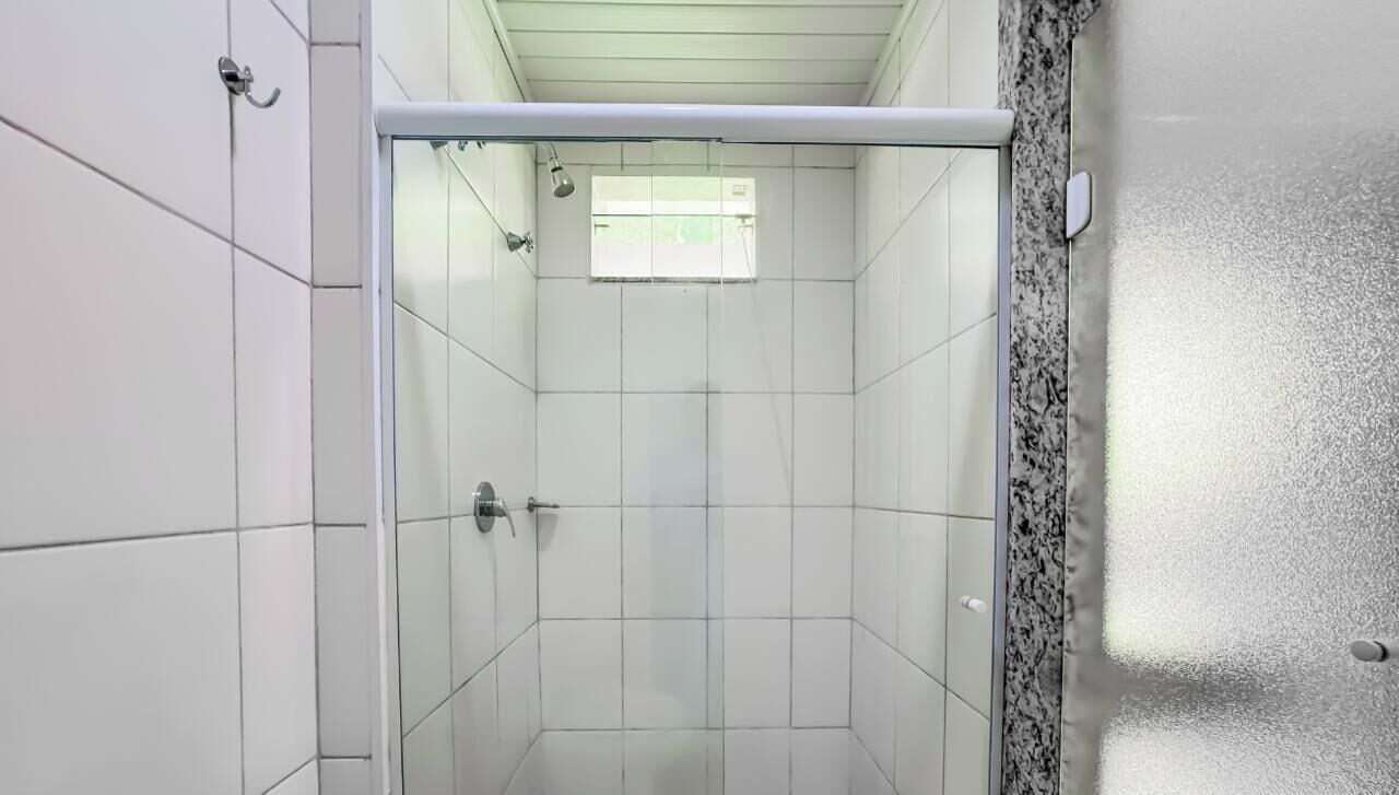 Ducha con azulejos blancos, puerta de cristal y una ventana pequeña.