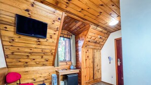 A wooden cabin interior with a TV mounted on the wall, a window with shutters, a small table with a kettle, and a door.