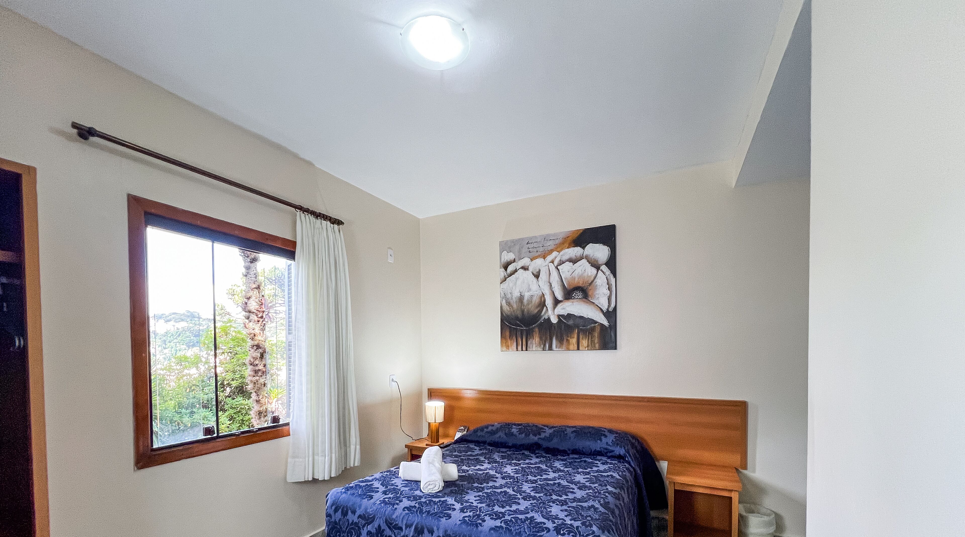 A bedroom with a bed, a nightstand, a lamp, a window with curtains, and a ceiling light.